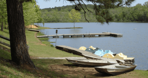 A serene lake scene with wooden boats, a dock, and lush green trees under a clear blue sky.