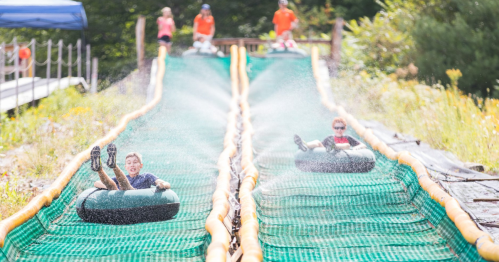 Two children slide down a water slide on inflatable tubes, with water spraying around them and adults supervising in the background.