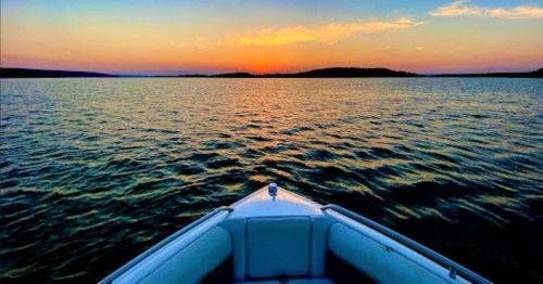 A boat's bow facing a tranquil lake at sunset, with vibrant colors reflecting on the water.