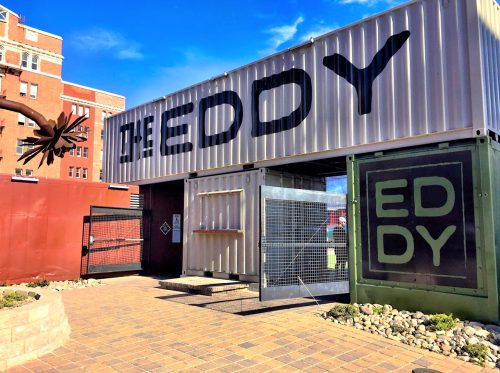Entrance to "The Eddy," a modern building made from shipping containers, with a blue sky in the background.