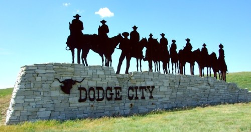 Silhouette of cowboys on horseback atop a stone sign reading "Dodge City" against a clear blue sky.