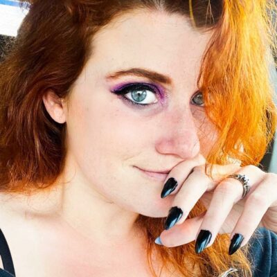 A close-up of a person with vibrant red hair and striking makeup, showcasing long black nails and a playful expression.