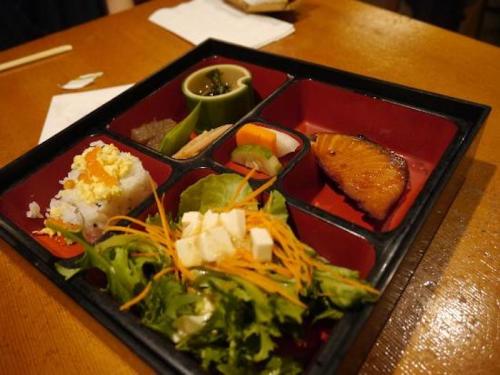 A bento box featuring sushi, salad, pickled vegetables, and grilled fish on a wooden table.