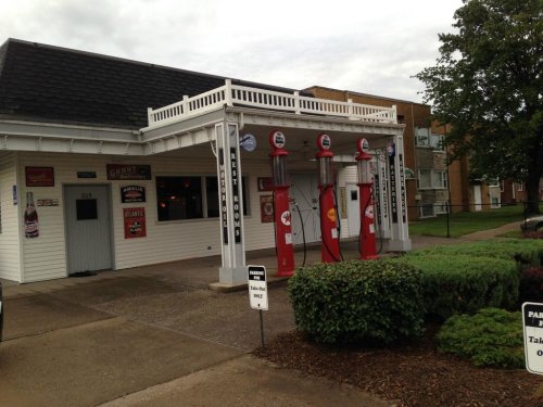 A vintage gas station with red pumps, white building, and retro signage, surrounded by greenery and parking signs.
