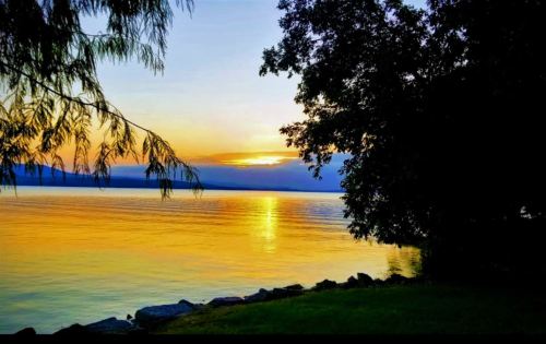 Sunset over a calm lake, with vibrant orange and yellow hues reflecting on the water, framed by trees and rocks.