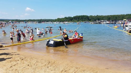 A busy beach scene with people swimming and relaxing, while a person paddles a colorful boat in the water.