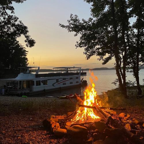 A cozy campfire in the foreground with a houseboat and sunset over the water in the background. Trees frame the scene.