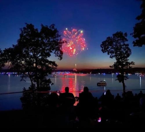 Fireworks illuminate the night sky over a lake, with silhouettes of people watching from the shore.