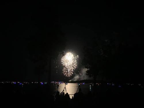 Fireworks illuminate the night sky over a lake, with silhouettes of people watching from the shore.