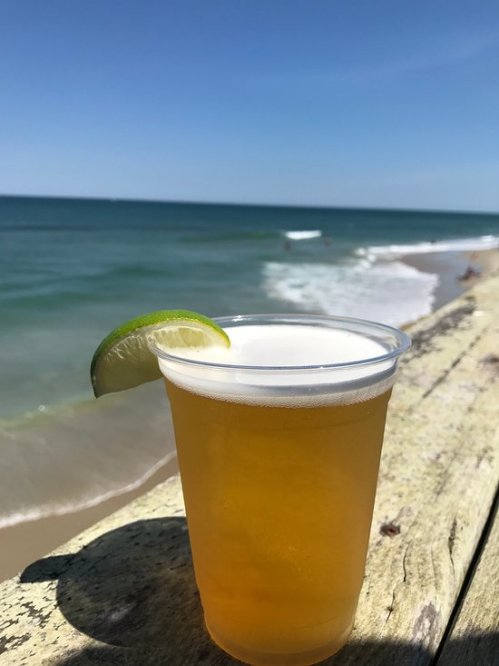 A cold beer in a plastic cup with a lime wedge, set against a sunny beach and ocean backdrop.