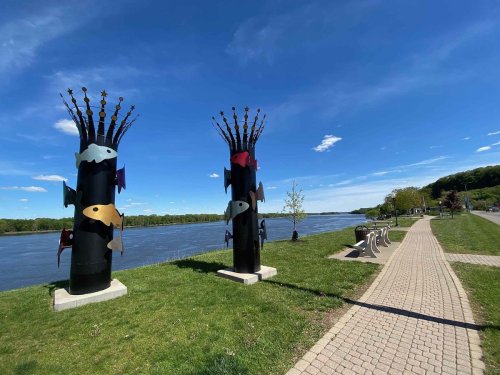 Two tall, colorful fish sculptures stand by a river, with a paved path and benches nearby under a clear blue sky.
