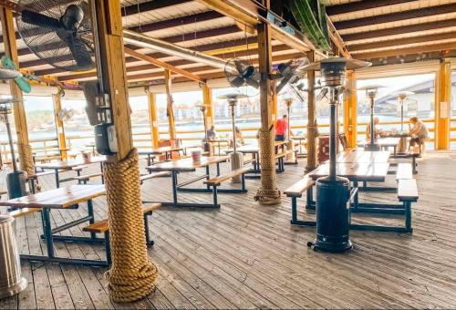A spacious outdoor dining area with wooden tables, fans, and a view of the water, partially filled with patrons.