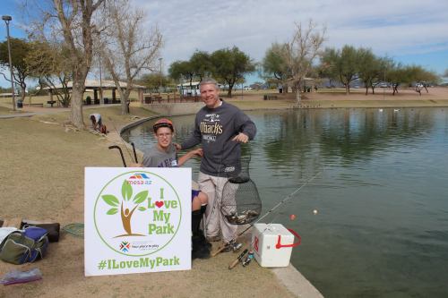A man and a boy smile by a pond, holding a fishing net, with a sign that reads "I Love My Park."