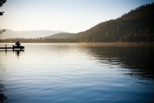 A serene lake at sunset, with a dock and two people silhouetted against the calm water and forested hills.