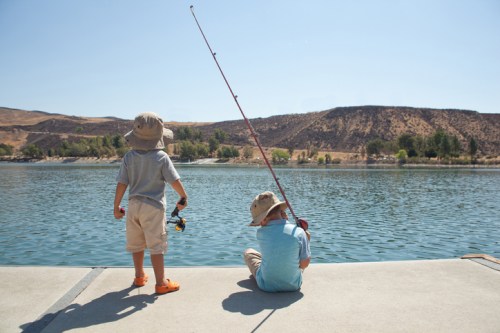 Two children fishing from a dock on a sunny day, with a calm lake and hills in the background.