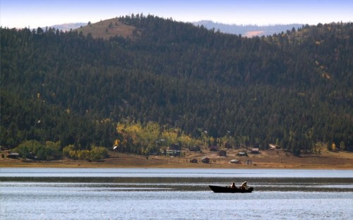 A small boat with two people glides across a calm lake, surrounded by forested hills and distant cabins.