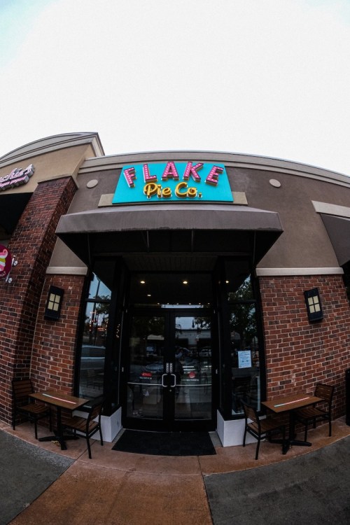 Exterior of Flake Pie Co. with a colorful sign, featuring a glass door and outdoor seating.