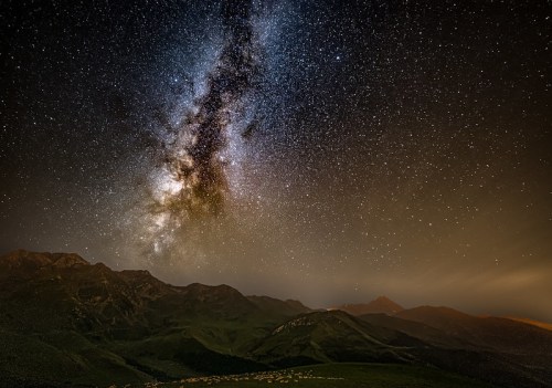 A stunning night sky filled with stars and the Milky Way over dark mountains and a faint horizon glow.