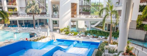 A vibrant indoor water park featuring a wave pool, palm trees, and lounge chairs in a modern setting.