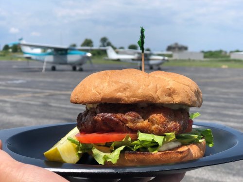 A hand holding a burger with lettuce, tomato, and pickle, with small planes in the background on an airstrip.