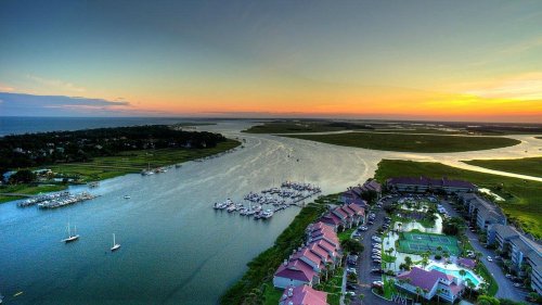 Aerial view of a serene river at sunset, with boats docked and lush greenery surrounding a waterfront community.