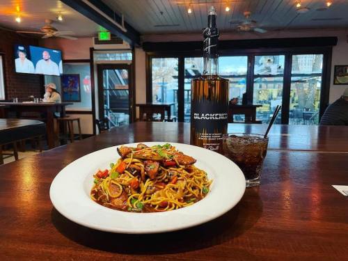 A plate of stir-fried noodles with vegetables and chicken, alongside a whiskey bottle and a glass of cola in a restaurant.