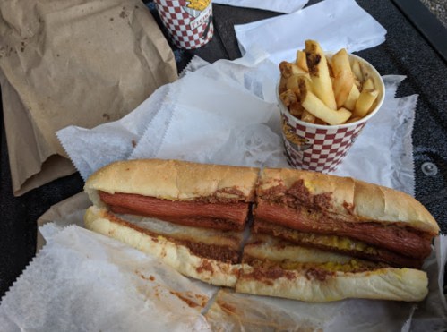 A hot dog in a bun with chili and mustard, accompanied by a cup of fries on a table with paper wrappers.