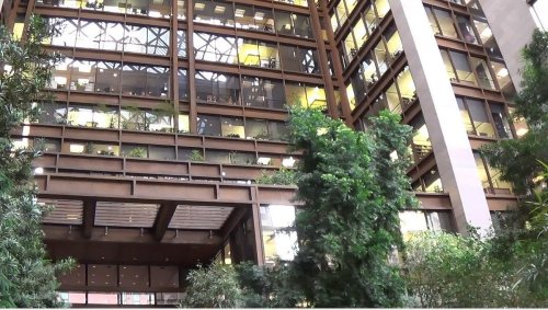 A modern building with large windows, greenery in the foreground, and multiple floors visible.