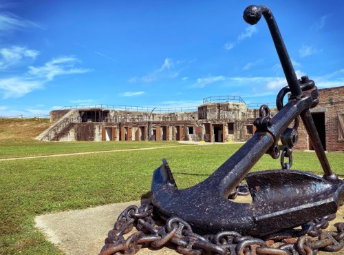 A large anchor in the foreground with a historic fort and blue sky in the background. Green grass surrounds the area.