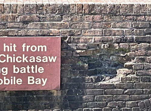 A weathered brick wall with a sign about damage from the Chickasaw during the Mobile Bay battle.