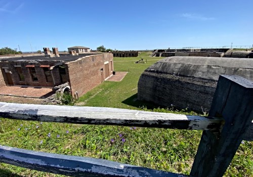 Historic fort buildings with grassy grounds and a clear blue sky in the background, viewed from a wooden fence.
