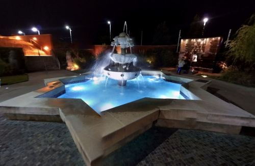 A star-shaped fountain illuminated at night, with water cascading into a blue-lit pool. Surrounding greenery and lights visible.
