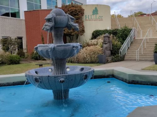 A decorative fountain with three tiers, surrounded by greenery and a statue, in front of a building labeled "H.U. Lee."