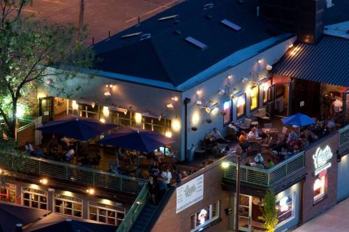 A lively outdoor restaurant with blue umbrellas, illuminated by warm lights, and patrons enjoying their meals.