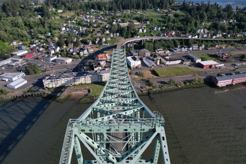 Aerial view of a green bridge curving over a river, with a small town and lush greenery in the background.