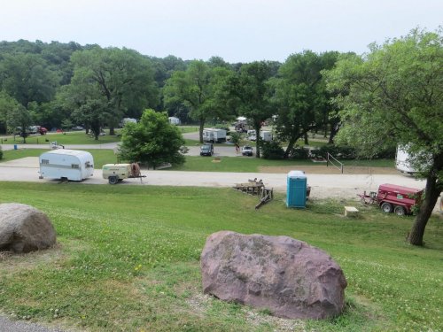 A scenic view of a campground with trailers, trees, and a portable restroom, set in a grassy area.