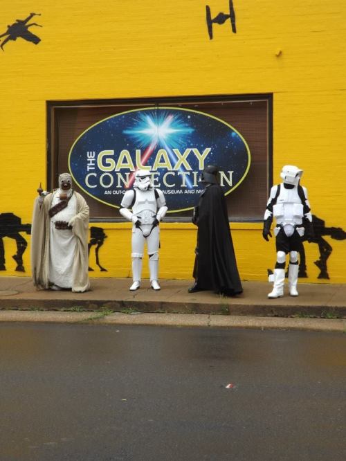 Four costumed characters, including a stormtrooper and a figure in a black cape, stand outside "The Galaxy Connection" store.