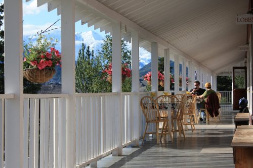 A serene porch with wooden chairs, flowers, and a view of mountains in the background, where two people are seated.