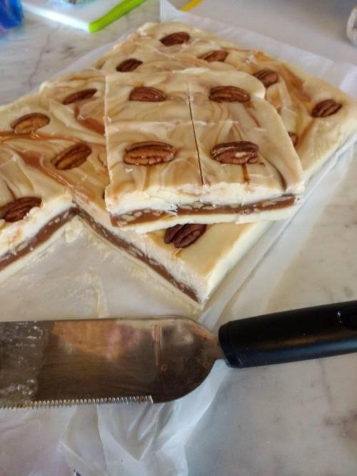 A close-up of layered fudge with pecans on top, cut into squares, with a knife resting beside it on a marble surface.