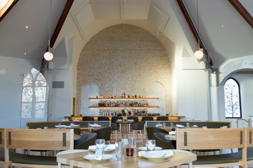 Modern restaurant interior with wooden furniture, a stone wall, and a well-stocked bar in a bright, airy space.