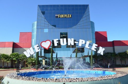 A large building with a sign reading "We Love Fun-Plex" and a fountain in front, under a clear blue sky.