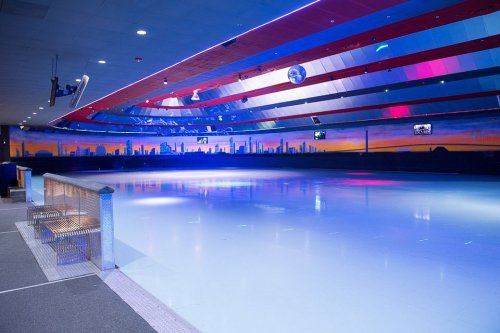 A brightly lit ice skating rink with colorful lights and a city skyline mural in the background.