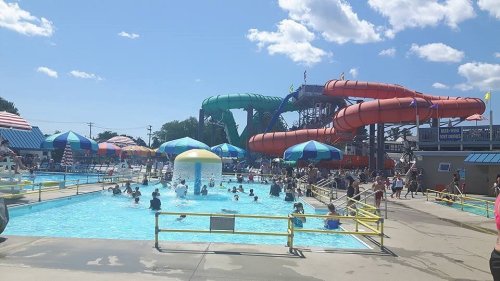 A busy water park with colorful slides, a pool filled with people, and umbrellas on a sunny day.