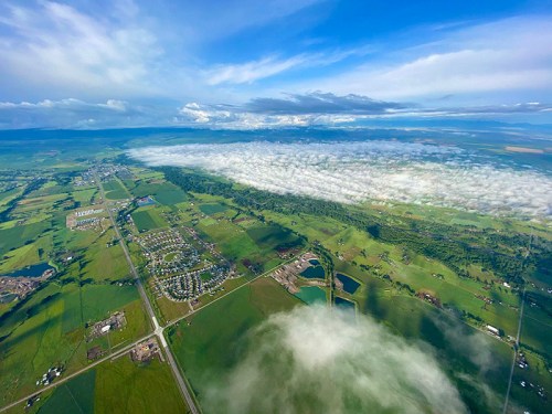 Aerial view of lush green fields and a town, with a blanket of clouds rolling over the landscape under a blue sky.