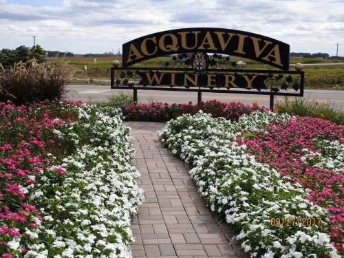 Sign for Acquaviva Winery surrounded by colorful flower beds and a paved pathway, with a scenic landscape in the background.