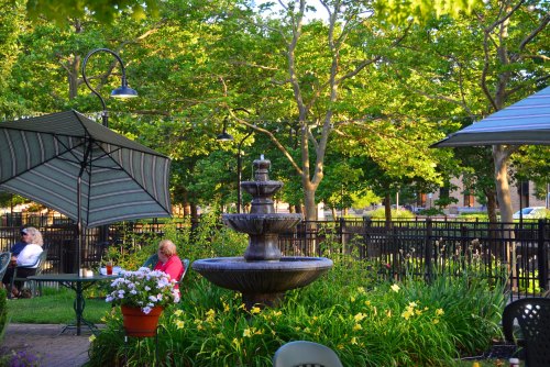 A peaceful garden scene featuring a fountain, lush greenery, and people sitting under umbrellas at a patio.