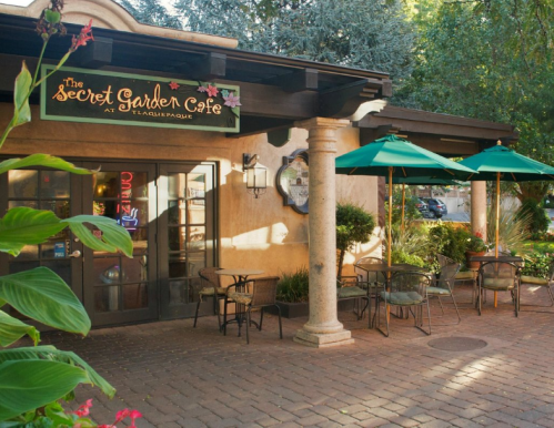 A charming café entrance with outdoor seating, green umbrellas, and a sign reading "The Secret Garden Café."