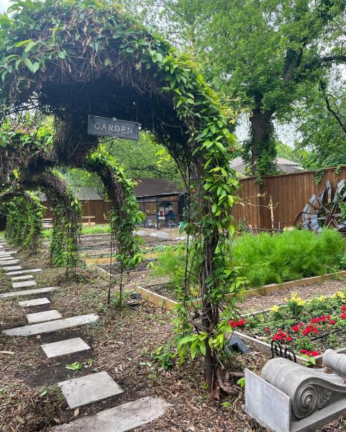 A garden path with stone steps leads under a leafy archway, featuring vibrant flowers and greenery in the background.
