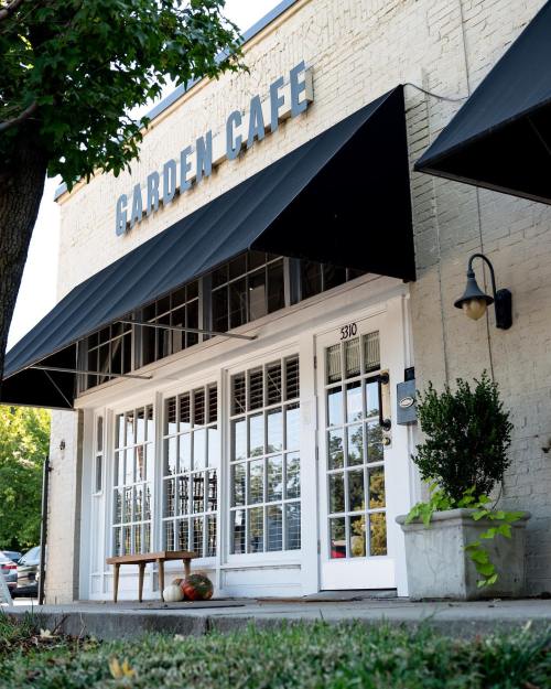 Exterior of Garden Cafe featuring large windows, a bench, and potted plants under a black awning.
