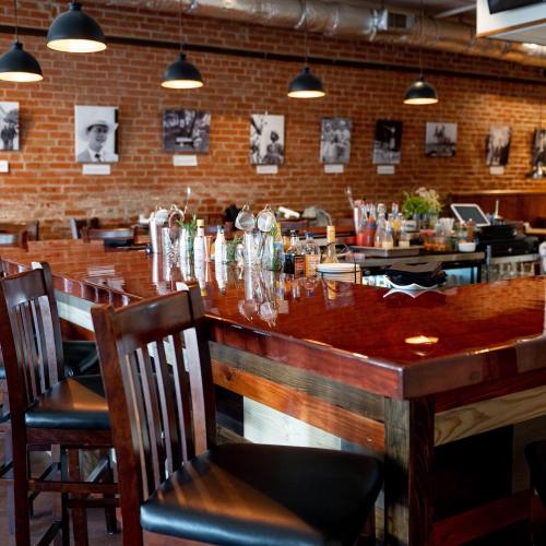 A cozy bar interior with wooden stools, a polished counter, and black-and-white photos on the brick wall.